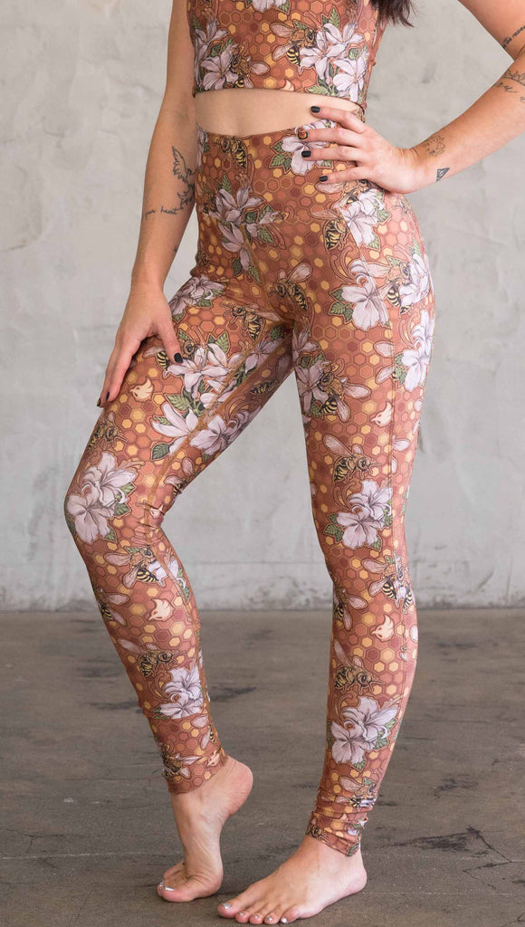 Front view of model wearing ultra lightweight "featherlight" leggings with clusters of honeybees and flowers. Her hand is in the side pocket that is big enough to hold a phone. 
