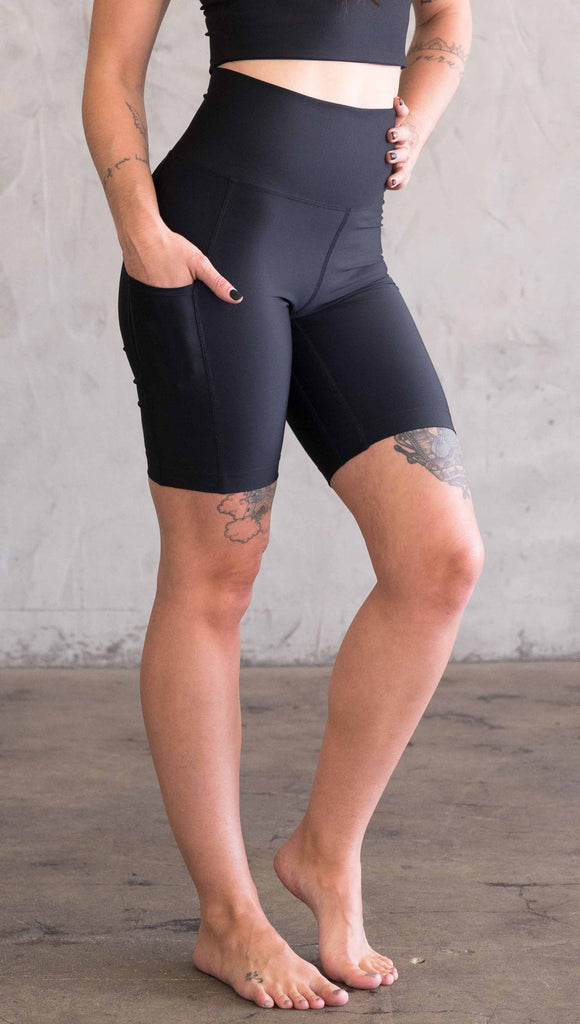 Waist down view of model wearing WERKSHOP bicycle length shorts in featherlight fabric. Black color with side cell phone pockets and a small eagle logo on the wearers left side