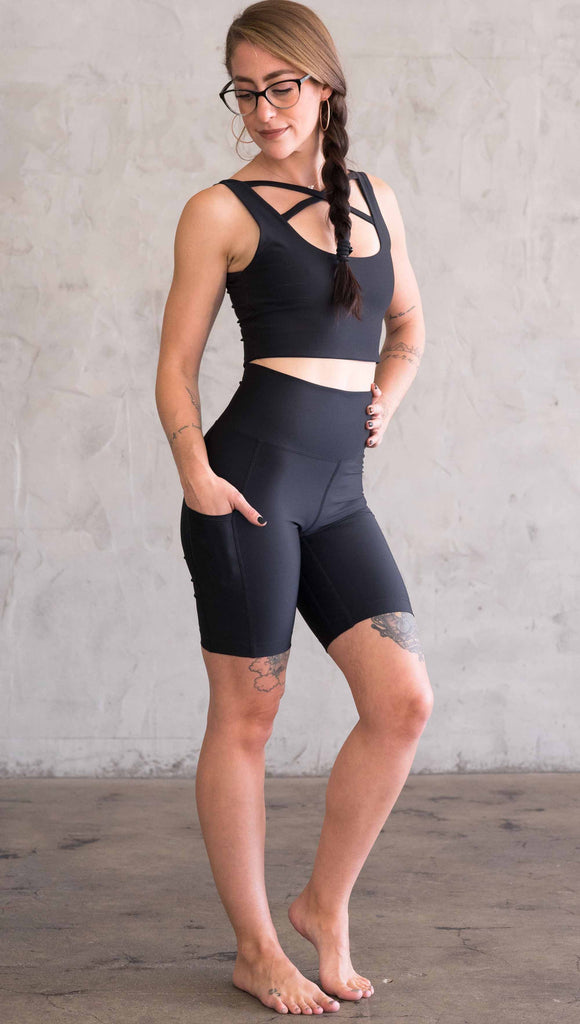 Full body view of model wearing WERKSHOP bicycle length shorts in featherlight fabric. Black color with side cell phone pockets and a small eagle logo on the wearers left side