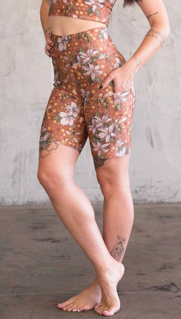Waist down side view of model wearing WERKSHOP Honeybees Bicycle Length Shorts. The artwork on the shorts features clusters of honeysuckle flowers and honeybees with a honeycmb background. Yellows, Coral, Orange with little pops of green.