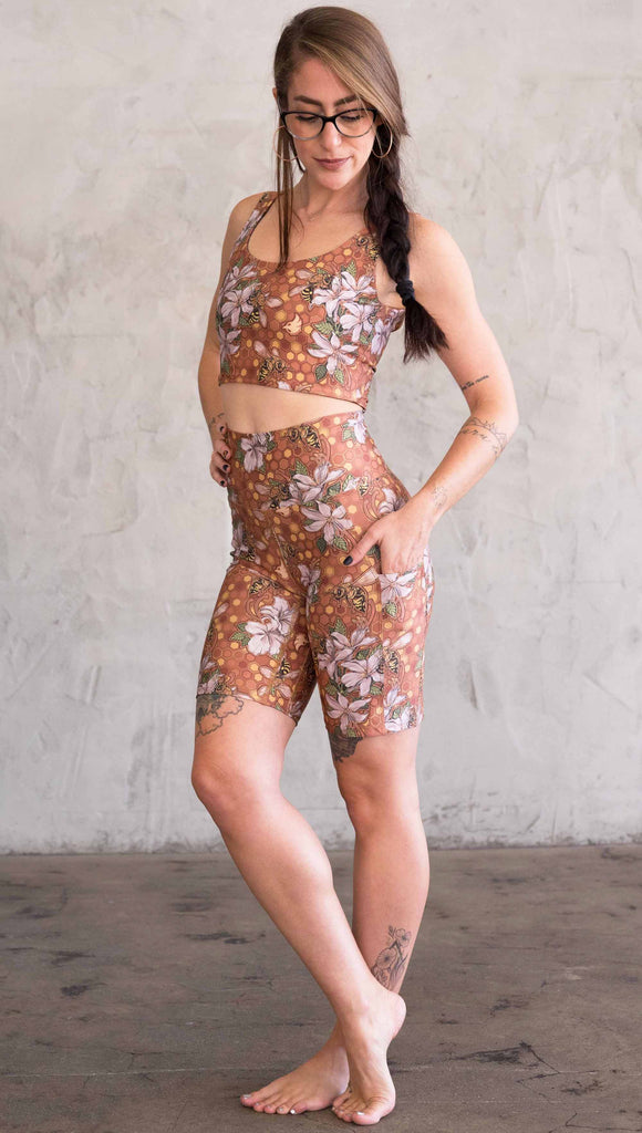 Full body font/side view of model wearing WERKSHOP Honeybees Bicycle Length Shorts. The artwork on the shorts features clusters of honeysuckle flowers and honeybees with a honeycmb background. Yellows, Coral, Orange with little pops of green.