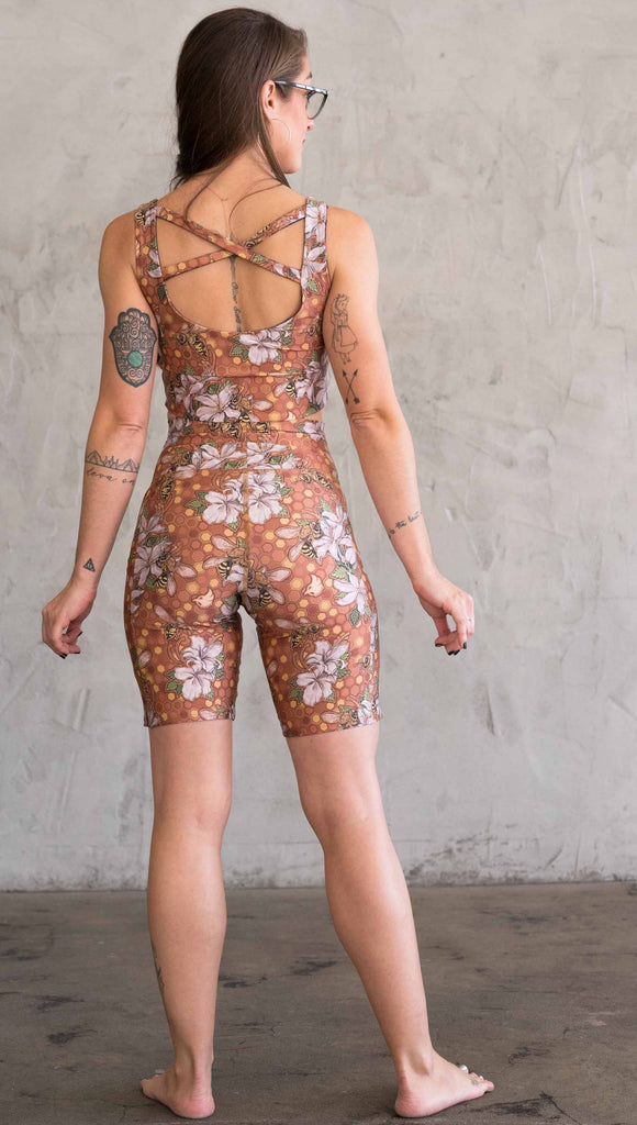 Full body back view of model wearing WERKSHOP Honeybees Bicycle Length Shorts. The artwork on the shorts features clusters of honeysuckle flowers and honeybees with a honeycmb background. Yellows, Coral, Orange with little pops of green.