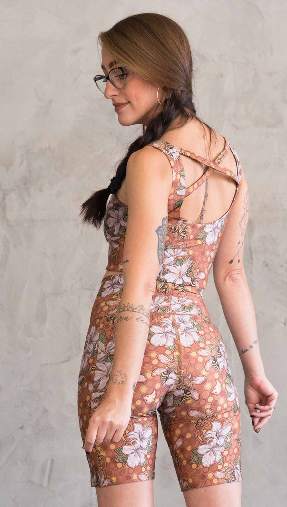 Zoomed in back view of model wearing WERKSHOP Honeybees Bicycle Length Shorts. The artwork on the shorts features clusters of honeysuckle flowers and honeybees with a honeycmb background. Yellows, Coral, Orange with little pops of green.