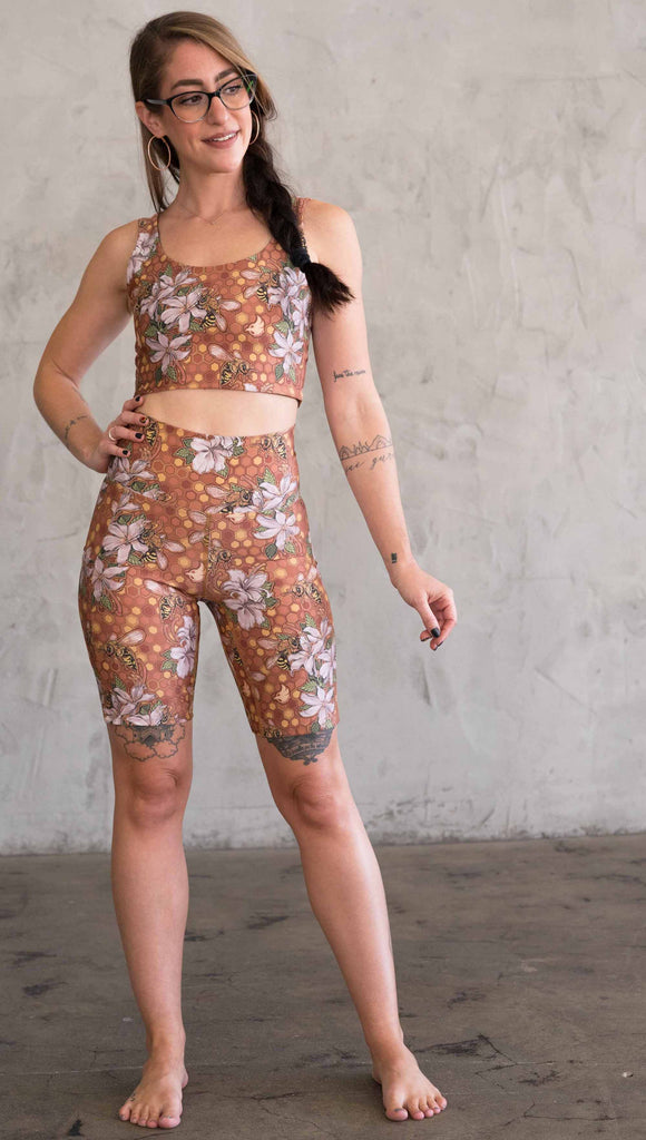 Full body front view of model wearing WERKSHOP Honeybees Bicycle Length Shorts. The artwork on the shorts features clusters of honeysuckle flowers and honeybees with a honeycmb background. Yellows, Coral, Orange with little pops of green.