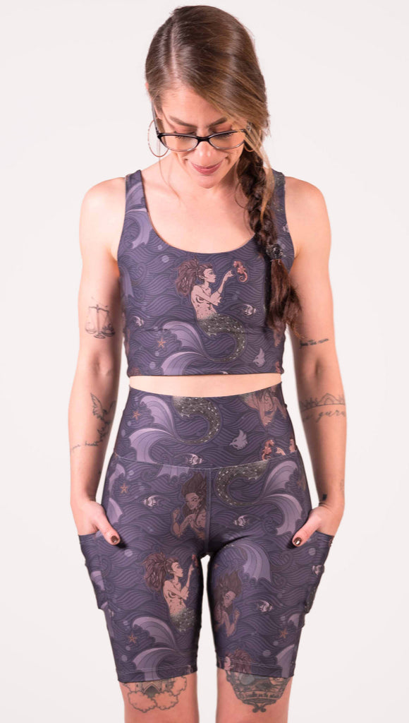 Zoomed in front view of model wearing WERKSHOP featherlight bicycle length shorts with Original Mermaids artwork. The mermaids have small intricate details on the fins and are swimming with seahorses and angel fish over a dark blue background with waves. The shorts have large pockets on each hip large enough to hold a phone.