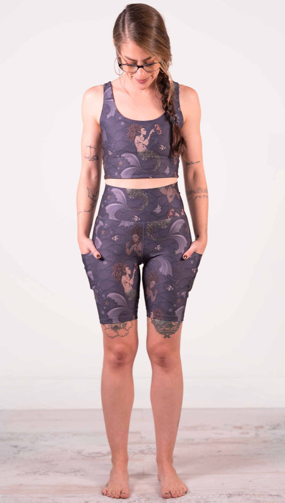 Full body front view of model wearing WERKSHOP featherlight bicycle length shorts with Original Mermaids artwork. The mermaids have small intricate details on the fins and are swimming with seahorses and angel fish over a dark blue background with waves. The shorts have large pockets on each hip large enough to hold a phone.