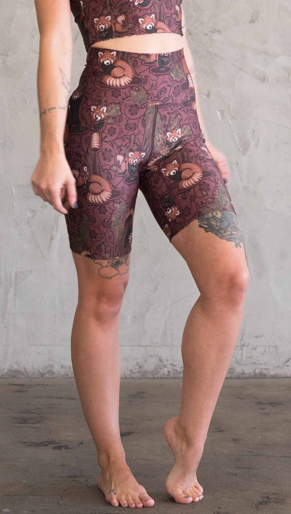 Waist down side view of model wearing WERKSHOP Red Panda Bicycle Length shorts. The artwork on the shorts features adorable little red pandas playing an having snacks next to abstract trees on a burgundy background.