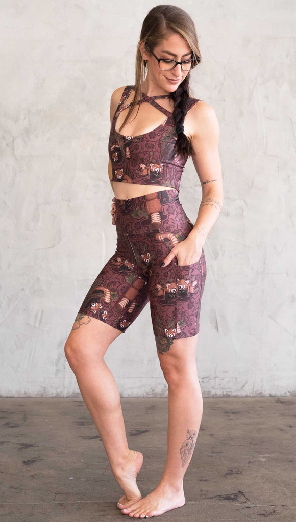 Full body side view of model wearing WERKSHOP Red Panda Bicycle Length shorts with her hand in her pocket. The artwork on the shorts features adorable little red pandas playing an having snacks next to abstract trees on a burgundy background.