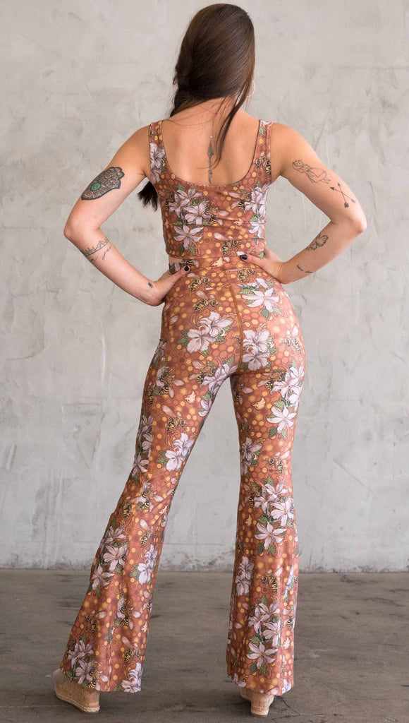 Full body back view of model wearing WERKSHOP Honeybee Bell Bottoms. The leggings have clusters of honeybees and flowers and phone pockets on each leg