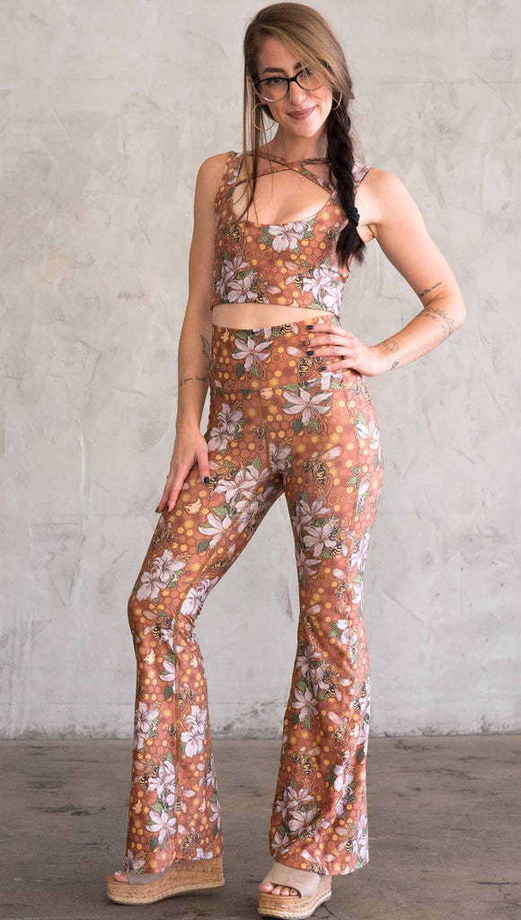 Full body front view of model wearing WERKSHOP Honeybee Bell Bottoms. The leggings have clusters of honeybees and flowers and phone pockets on each leg
