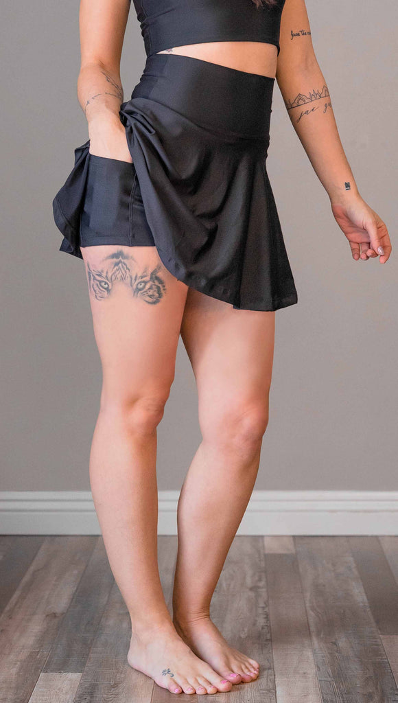Waist down view of model wearing WERKSHOP Tennis Skirt, with the skirt lifted up to show the shorts and pockets underneath. In solid black color