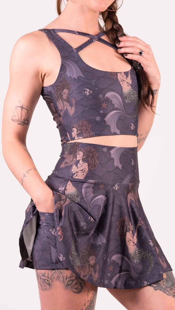 Zoomed in view of model wearing WERKSHOP featherlight skirt with Original Mermaids artwork. The mermaids have small intricate details on the fins and are swimming with seahorses and angel fish over a dark blue background with waves. The skirt has built-in shorts underneath with side pockets big enough to hold a cellphone.