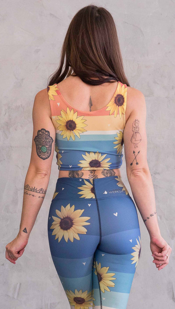 Back view of model wearing WERKSHOP Sunflower Rainbow Top. The top has vintage colored rainbow stripes on both sides. The stripes start orange at the shoulders, yellow and cream across the chest and then blue at the sweep. One side also has sunflowers and small cream colored hearts.