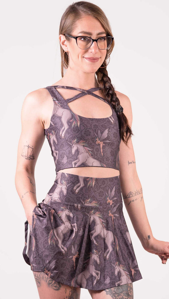 Front view of model wearing WERKSHOP Four-Way Reversible Top with Original Mermaids artwork on one side and Unicorn artwork on the opposite side. She is shown wearing the unicorns on the outside with the “X” strap detail in the front. The unicorns have soft rainbow-colored hair with a pixie friend over a purple background.