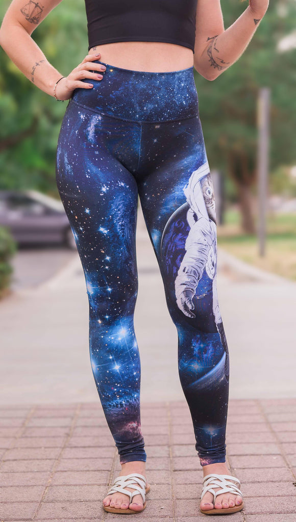 Model wearing Catstronaut Leggings. The leggings are printed with a cat in an astronaut suit on the wearer's left leg. The galaxy background is a deep cobalt blue with nebula swirls and shooting stars.
