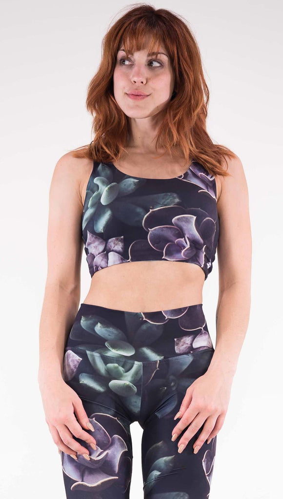 Front view of model wearing the Midnight Garden reversible top. This is in the Midnight Garden side, it is a black crop top with green and purple succulent plants throughout