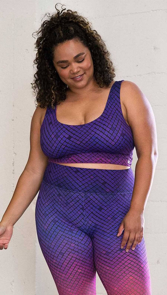 Three quarter length front view of model wearing a reversible crop top with a purple mosaic print on one side and solid fuschia with a small white Eagle logo on the other side