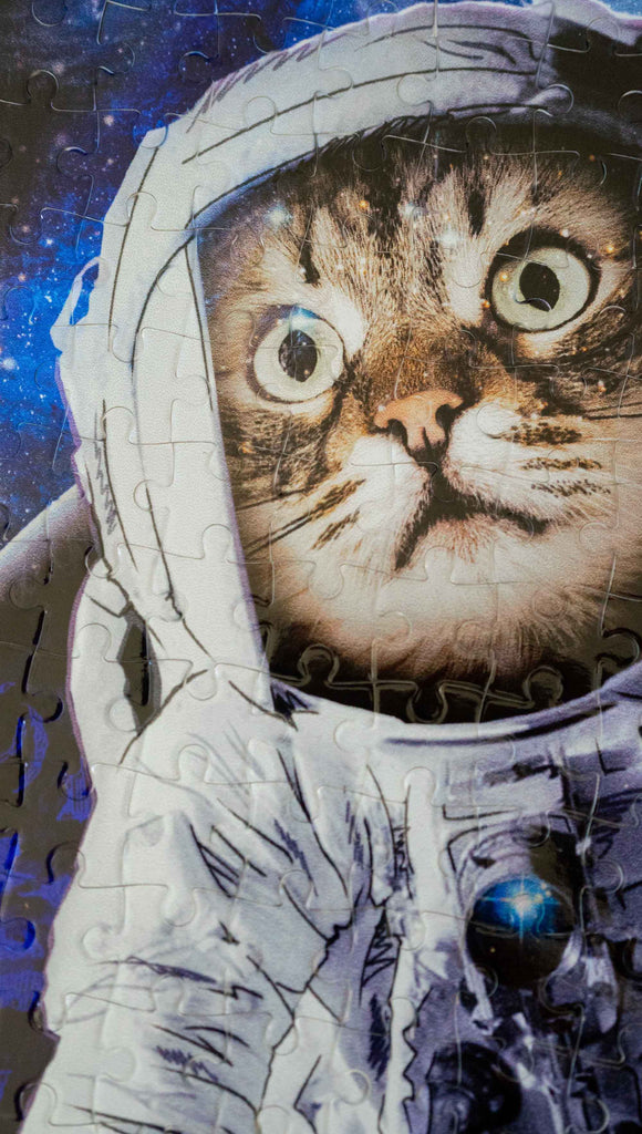 Zoomed in view WERKSHOP Space Cat (Catstronaut) Puzzle. The artwork features a house cat wearing an astronaut uniform, floating in outer space with a nebula behind him.