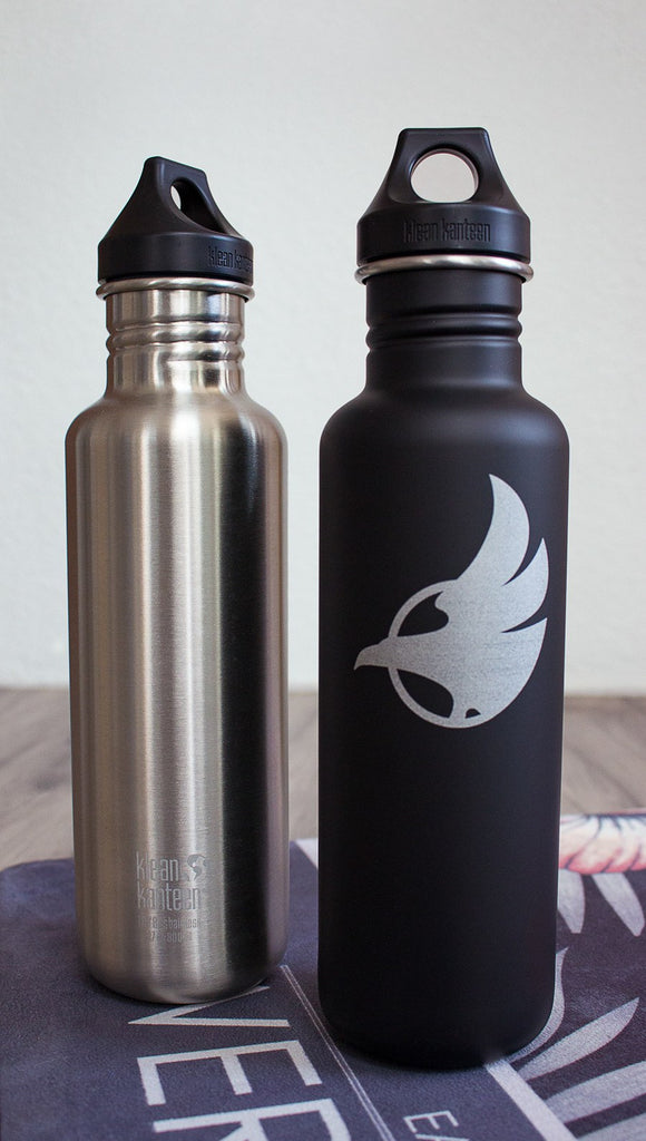 Black water bottle with Eagle Rock Werkshop logo next to silver water bottle with Eagle Rock Werkshop logo resting on a purple yoga mat with Eagle Rock Werkshop logo.