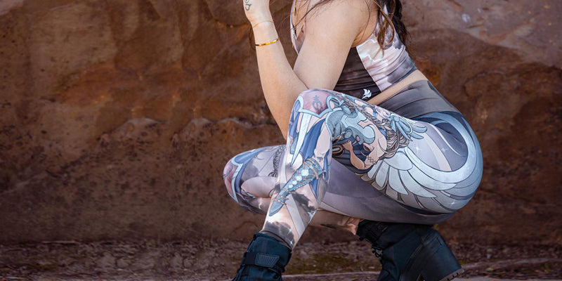 Model casually squatting while wearing WERKSHOP Valkyrie leggings. The leggings are printed with original artwork by Chriztina Marie and feature a Valkyrie warrior from Norse Mythology flying through a dark sky holding a shield and two swords.