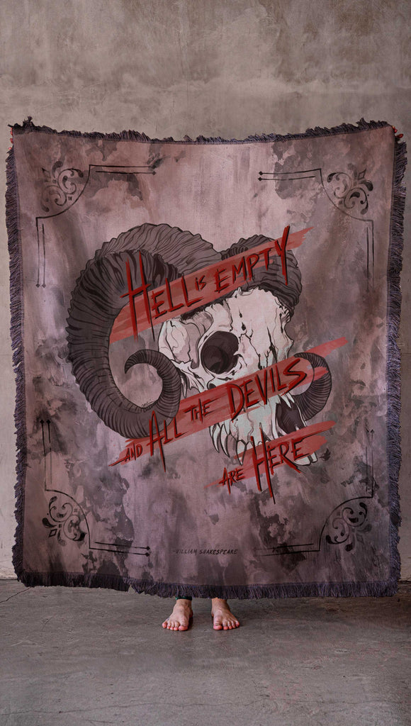 Person holding a large decorative blanket with a skull and text design against a concrete wall. Featuring the quote, "Hell is Empty and All The Devils Are Here" by William Shakespeare from the play, The Temptest.