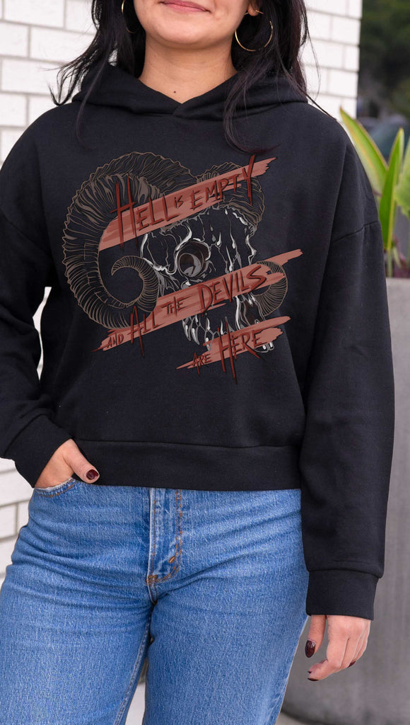 Person wearing a black hoodie with graphic design and text, standing against a white wall. Featuring a gothic skull and the quote, "Hell is Empty and the Devils Are All Here" from the Temptest by William Shakespeare