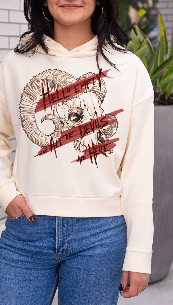 Person wearing a cream color hoodie with a graphic design of a ram and text, standing outdoors. Featuring a gothic skull and the quote, "Hell is Empty and the Devils Are All Here" from the Temptest by William Shakespeare.