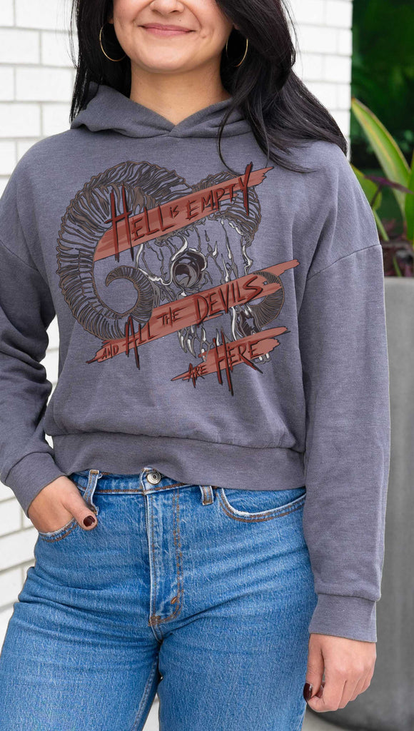 Person wearing a gray hoodie with graphic design and text, standing outdoors. Featuring a gothic skull and the quote, "Hell is Empty and the Devils Are All Here" from the Temptest by William Shakespeare
