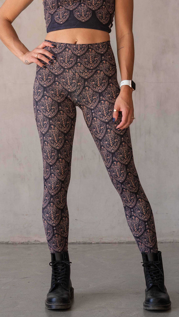 Front view of model wearing WERKSHOP Dragon Rider leggings in Gold. The artwork features an intricate battle shield designed to look like dragon scales. This color way is all shades of gold and bronze.