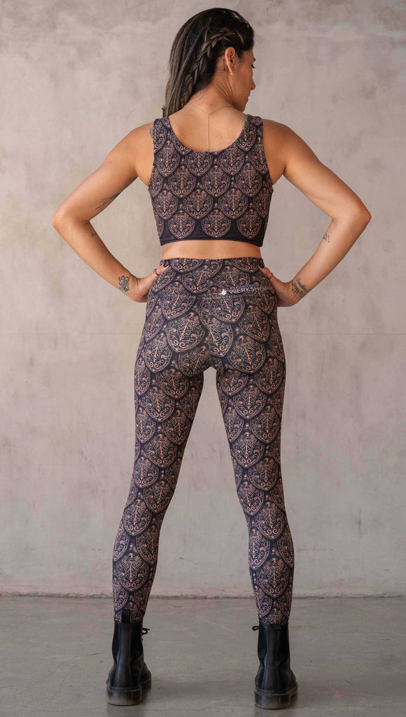 Full body back view of model wearing WERKSHOP Dragon Rider leggings in Gold. The artwork features an intricate battle shield designed to look like dragon scales. This color way is all shades of gold and bronze.