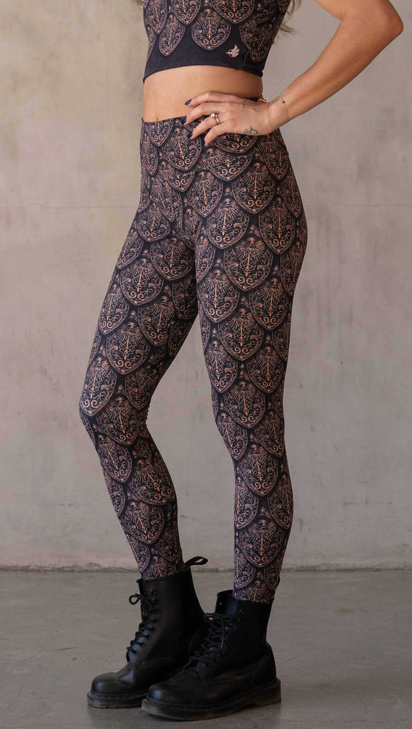 Side view of model wearing WERKSHOP Dragon Rider leggings in Gold. The artwork features an intricate battle shield designed to look like dragon scales. This color way is all shades of gold and bronze.