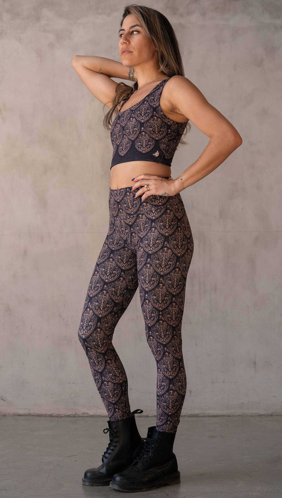 Full body side view of model wearing WERKSHOP Dragon Rider leggings in Gold. The artwork features an intricate battle shield designed to look like dragon scales. This color way is all shades of gold and bronze.