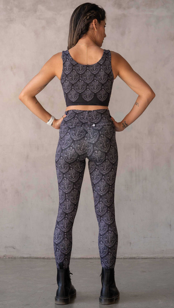 Full body back view of model wearing WERKSHOP Dragon Rider leggings in Silver. The artwork features an intricate battle shield designed to look like dragon scales. This color way is all shades of black and gray.