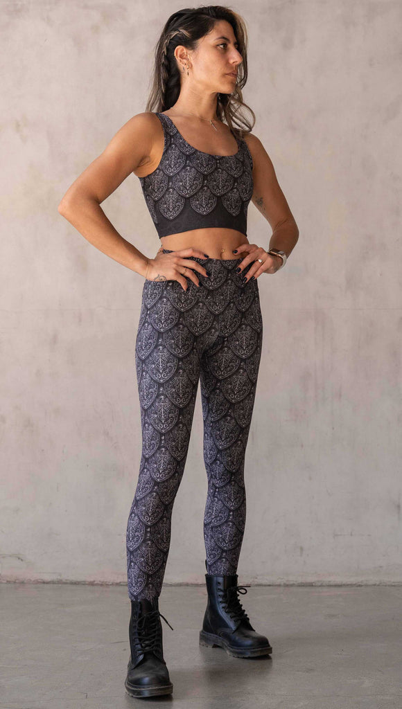 Full body side view of model wearing WERKSHOP Dragon Rider leggings in Silver. The artwork features an intricate battle shield designed to look like dragon scales. This color way is all shades of black and gray.