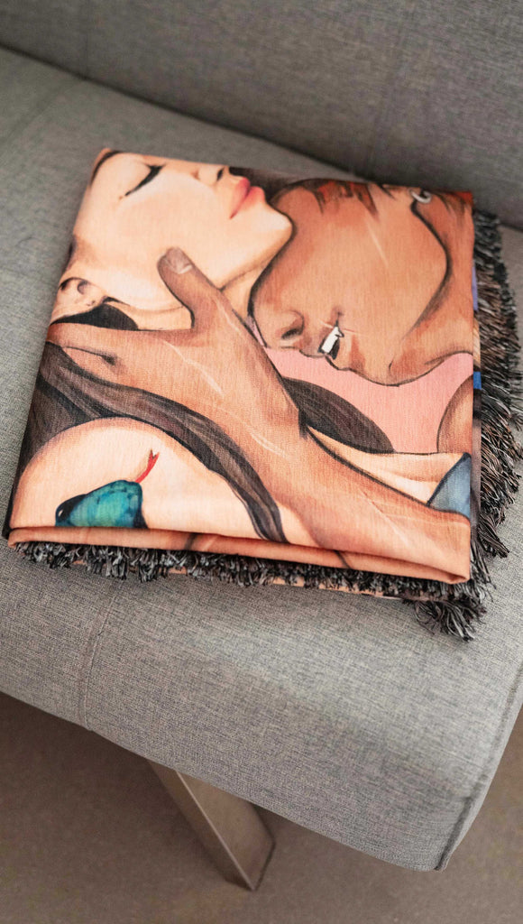 Officially Licensed WERKSHOP X The Ever King Tapestry/Blanket. "The Serpent and the Singbird" The artwork features Erik Bloodsinger passionately kissing Livia in front of a porthole window with a rope wrapping aroung it and the quote, "Hate me all you want, but don't regret me" - folded on a gray sofa