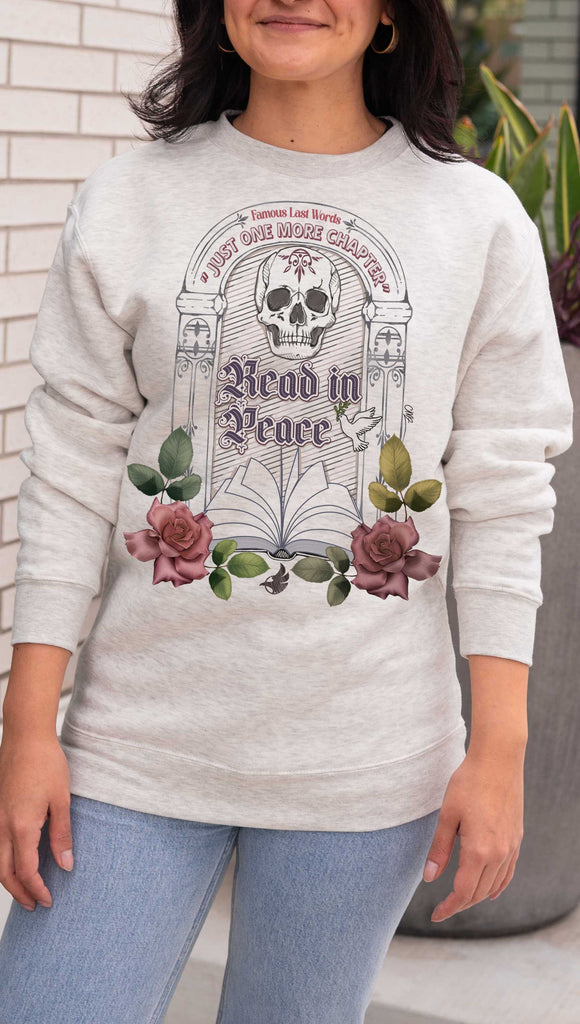 Person wearing a light gray (oatmeal) sweatshirt with a 'Read in Peace' design featuring a skull and flowers over a tombstone with the phrase, "Famous Last Words: Just One More Chapter" 