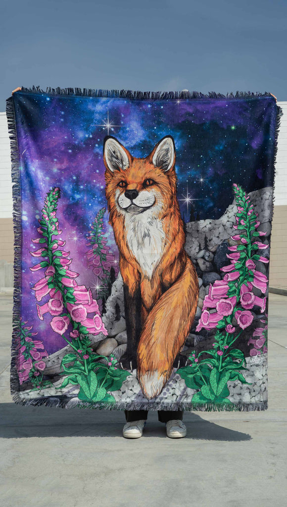 girl holding up WERKSHOP's Save A Fox Tapestry. The tapestry features soft chenille fabric, with original artwork of a fox in front of a galactic background with foxglove plants framing him.