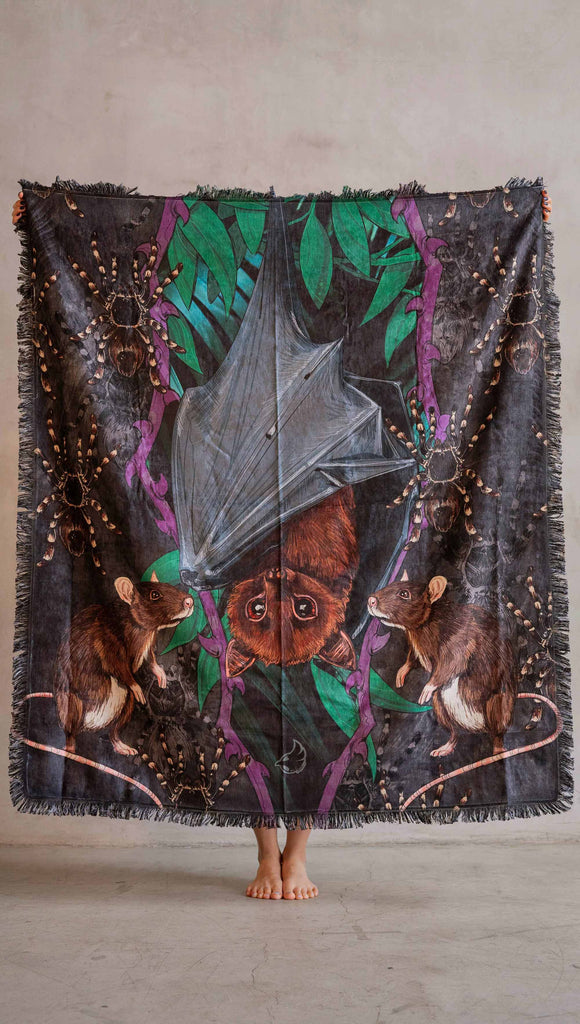 Decorative Chenille Tapestry printed with limited edition artwork by our Female Founder, Chriztina Marie. The artwork features an adorable fruit bat dangling upside down inside a tropical scene with a purple wreath of thorns. Under the bat, there are two rats. The background is a distressed dark gray brushstroke texture.