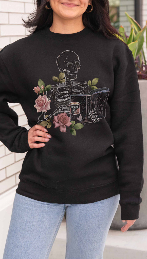 Person wearing a black sweatshirt with a skeleton and floral design, standing outdoors. The pullover features the bookish phrases, "Death by TBR" and "A Beginners Guide: How to Find Time to Read More Smut"