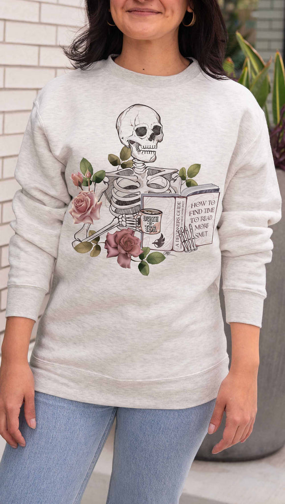 Person wearing a light gray (oatmeal) sweatshirt with a skeleton and floral design, standing outdoors. The pullover features the bookish phrases, "Death by TBR" and "A Beginners Guide: How to Find Time to Read More Smut"