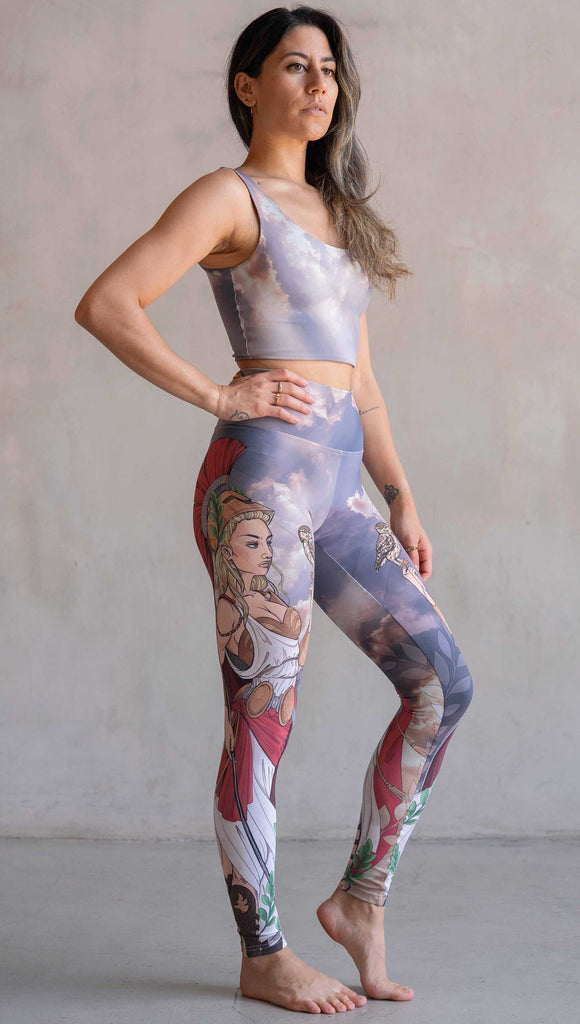 Full body view of model wearing WERKSHOP Athena leggings. The leggings are printed with original artwork by Chriztina Marie and features Athena, the goddess of war standing on a cliff’s edge. She is holding a spear with one hand and her owl with the other. She is wearing a greek goddess dress/warrior hat with colors of cream, gold and red. Behind her is a beautiful cloudy sky with rays of light shining down onto her. The model is also wearing a matching top.