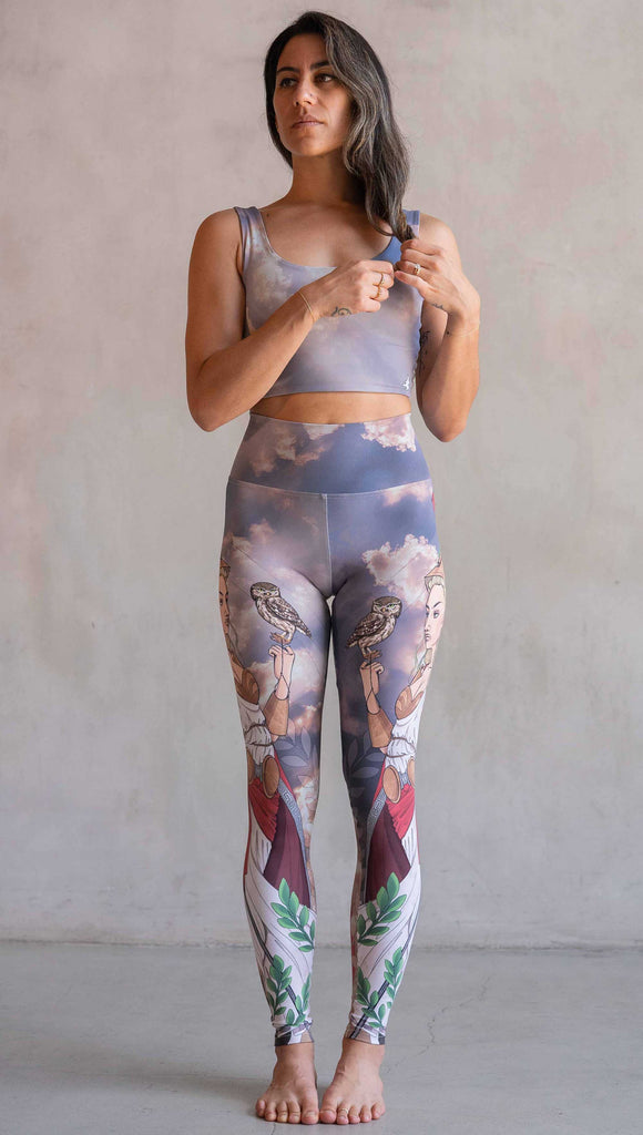 Full body front view of model wearing WERKSHOP Athena leggings. The leggings are printed with original artwork by Chriztina Marie and features Athena, the goddess of war standing on a cliff’s edge. She is holding a spear with one hand and her owl with the other. She is wearing a greek goddess dress/warrior hat with colors of cream, gold and red. Behind her is a beautiful cloudy sky with rays of light shining down onto her. The model is also wearing a matching top.