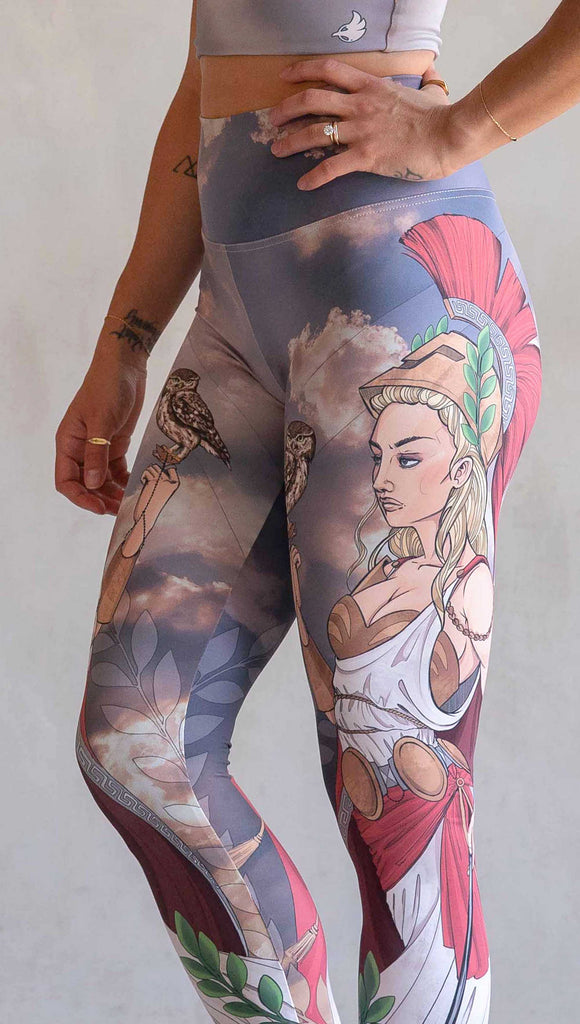 Zoomed in view of model wearing WERKSHOP Athena leggings. The leggings are printed with original artwork by Chriztina Marie and features Athena, the goddess of war standing on a cliff’s edge. She is holding a spear with one hand and her owl with the other. She is wearing a greek goddess dress/warrior hat with colors of cream, gold and red. Behind her is a beautiful cloudy sky with rays of light shining down onto her. The model is also wearing a matching top.