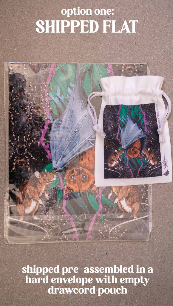 WERKSHOP Spooky Season jigsaw puzzle The artwork features an adorable fruit bat dangling upside down inside a tropical scene with a purple wreath of thorns. Option 1: Shipped pre-assembled in a hard envelope with an empty matching drawcord pouch.