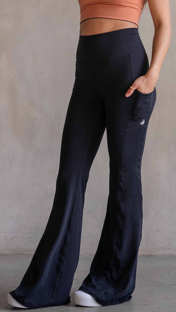 Model wearing WERKSHOP Black EnviSoft Bells. The pants feaure a belled leg opening, flatlock seams, pockets on each thigh, a long panel gusset and small silver eagle logo on the right pocket.