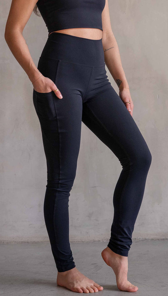 Model wearing WERKSHOP Solid Black EnviSoft Leggings. Featuring pockets on each thigh, a long panel gusset, flatlock seams and an eagle logo on the right pocket.