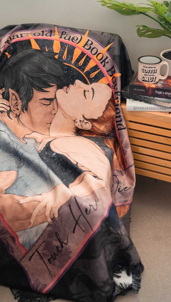 WERKSHOP soft chenille tapestry with a original and exclusive artwork of a fae man passionately kissing a woman. The phrase on the artwork says, "daydreaming of my 500 year old fae book boyfriend" on top and "touch her and die' on the bottom. the background is a distressed dark mauve with charcoal splatter. Draped over the edge of a gray sofa.