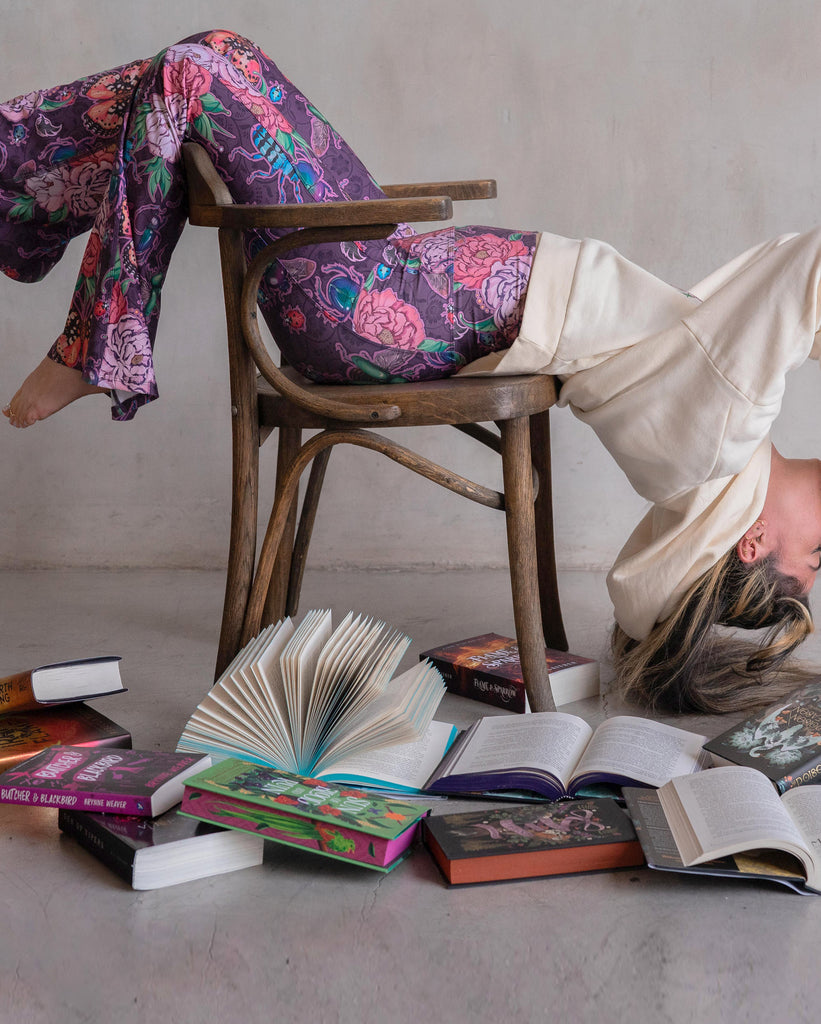 Girl doing a moderate backbend over a chair while reading her book. She is wearing WERKSHOP Enchanted Garden bells and "A Book A Day Keeps Reality Away" hoodie.