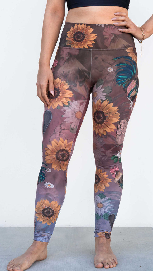 Model wearing WERKSHOP "Cocks n Beavers" Leggings. The leggings show hand painted artwork of roosters and beavers with daisies and sunflowers over an ombreback ground that goes from a warm brown on top to slate blue at the leg opening.