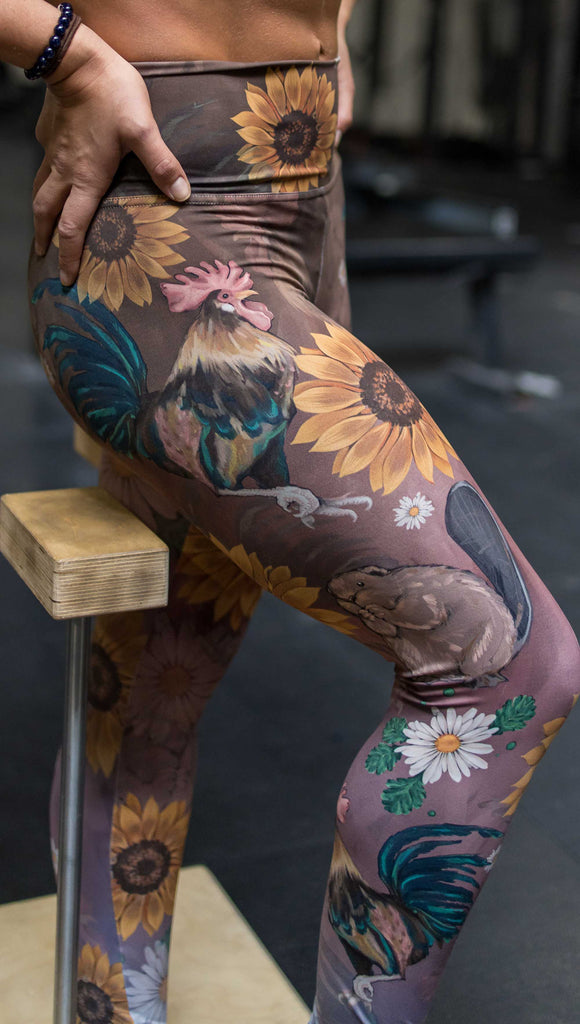 Model wearing WERKSHOP "Cocks n Beavers" Leggings. The leggings show hand painted artwork of roosters and beavers with daisies and sunflowers over an ombreback ground that goes from a warm brown on top to slate blue at the leg opening.